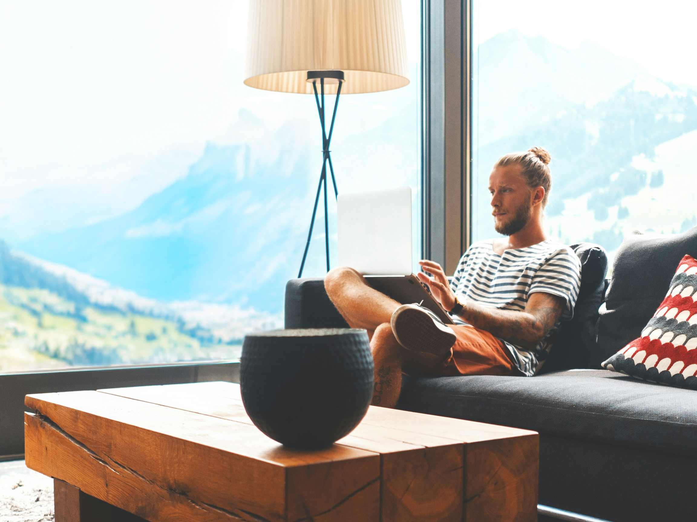 Header Bild Blogartikel Digitalinkasso in der Schweiz. Mann Mitte 30 sitzt auf einer Couch und hat einen Laptop auf dem Schoß. Im Hintergrund sieht man durch große Fenster die schweizer Berge.